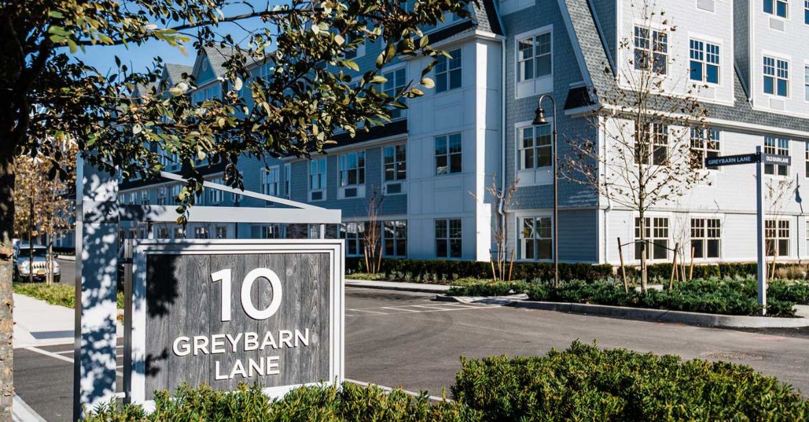 In the foreground, a grey sign with white lettering says "10 Greybarn Lane". In the background stands a three story complex, grey and white, with large windows and green landscaping in front.
