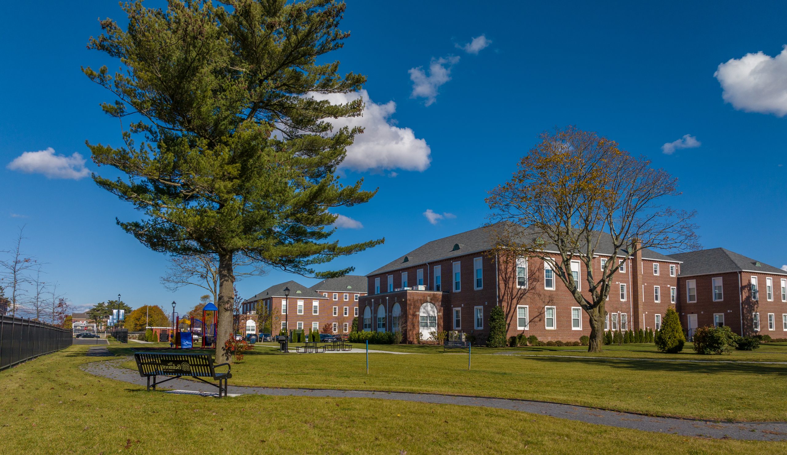 In the foreground is a large green lawn with trees, in the background is a red rental building with white trim.