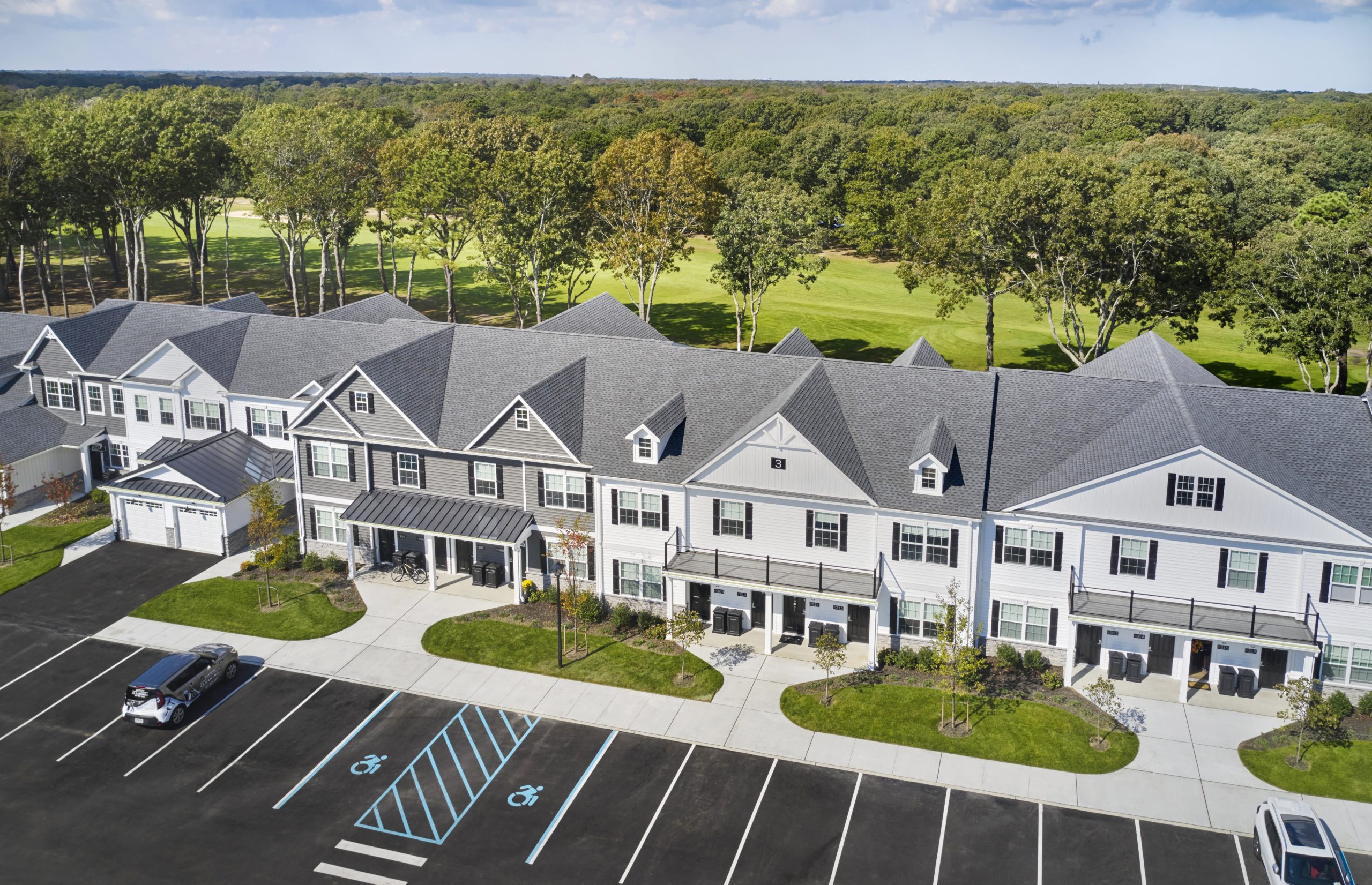 An aerial view of a white and grey complex of town homes. There are green lawns and parking spaces in front of the building, and green space with trees behind the building.